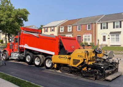 A red dump truck and yellow paver laying fresh asphalt on a residential street near townhouses.