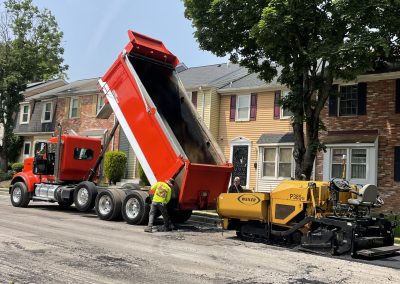 A red dump truck unloads asphalt into a paving machine on a residential street, with a worker nearby.