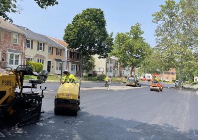 Workers use heavy machinery to pave a residential street with fresh asphalt on a sunny day.