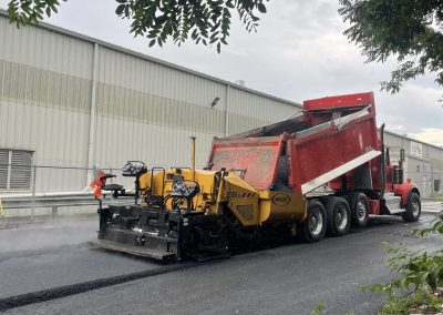 A red dump truck unloads asphalt into a yellow paver on a freshly paved road near an industrial building.