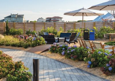 Outdoor patio with wooden chairs, umbrellas, flowers, and pathways on a sunny day with houses in the background.