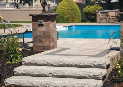 Three stone steps lead to a backyard patio with a pool, lamp post, and landscaped plants.