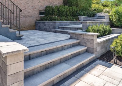 Stone steps and patio with a metal railing, surrounded by green shrubs and a brick house wall.