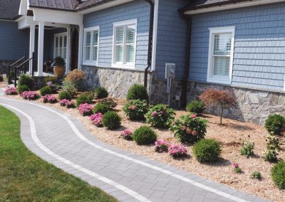 Curved stone walkway with flower beds and shrubs beside a blue house with stone accents.