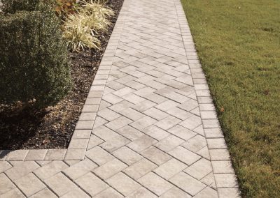 Stone paver walkway bordered by shrubs and grass beside a building, leading away in perspective.