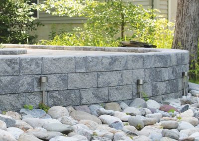 Gray stone retaining wall with metal lights, surrounded by smooth rocks and greenery in a sunny yard.