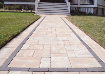 Wide stone walkway with dark border leading to steps and porch of a house with green lawn on both sides.