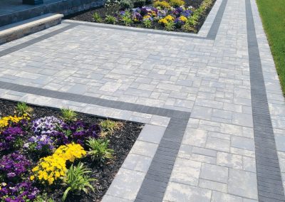 A paved walkway bordered by flower beds with colorful flowers and green grass near a building.