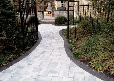 A curved stone walkway leads through a black metal gate to a patio with outdoor furniture and greenery.