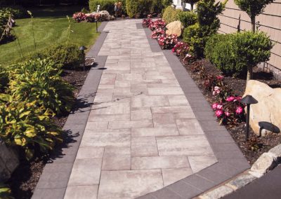 A stone walkway bordered by flowers, bushes, and garden lights leads to a house with trimmed shrubs.