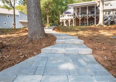 Gray stone walkway with steps leading uphill toward a house with a large porch, surrounded by trees.