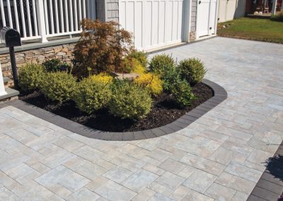 A small landscaped garden with shrubs and a tree, bordered by a stone patio near a house and white fence.