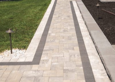 A stone paver walkway with a dark border runs between grass and a mulched garden bed with outdoor lighting.