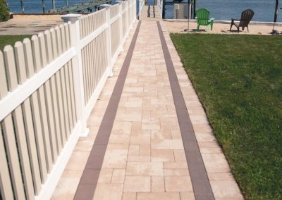 A paved walkway leads to a dock with a boat, bordered by a fence, grass, and a sandy area with chairs.