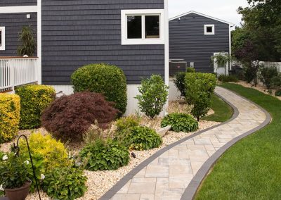 Curved stone walkway beside a landscaped garden with bushes, leading past a dark gray house.