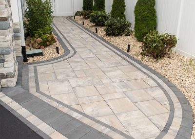 Stone walkway with decorative border, flanked by shrubs and white fence, leading to a backyard area.