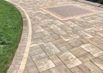 A paved patio with tan and gray tiles, bordered by grass and a metal fence, with a brick square inset.