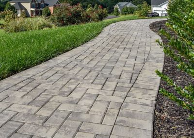 Curved stone paver walkway through landscaped grass and bushes in a suburban neighborhood.
