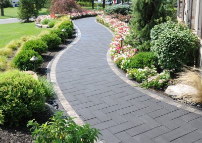 Curved stone pathway bordered by lush green bushes and colorful flowers in a landscaped garden.