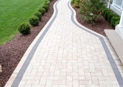 Curved stone walkway bordered by dark bricks, shrubs, and mulch in a well-kept front yard.