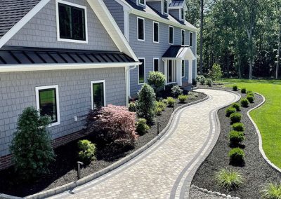 Curved stone walkway with landscaped bushes and trees leading to a large grey house with white trim.