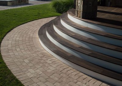 Curved wooden steps lead down to a patio with brick pavers, surrounded by green grass and outdoor seating.