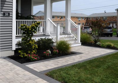 Front porch with white railing, landscaped garden, and paved walkway beside a green lawn in a suburban neighborhood.