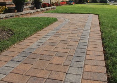 Curved stone paver walkway bordered by grass leading to steps and a row of houses.