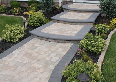 Stone walkway with curved steps, surrounded by green shrubs, flowers, and garden lights leading to a house.