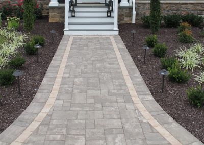 Stone walkway with light border leads to white stairs and front porch, surrounded by landscaped plants and mulch.