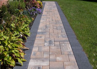 A paved garden walkway with gray and tan bricks, bordered by flowers on one side and grass on the other.