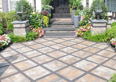 Front patio with checkerboard pavers, lush green plants, and colorful flowers near a house entrance.