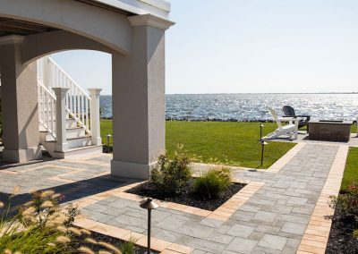 Paved patio with garden beds, stairs, and chairs facing a waterfront view under a clear sky.