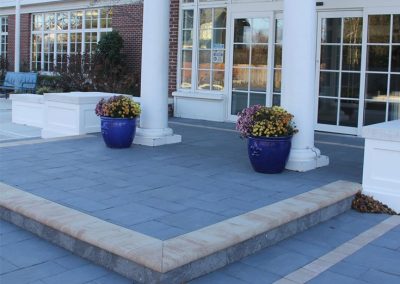 Blue planters with flowers on a paved entrance with columns and large windows outside a building.