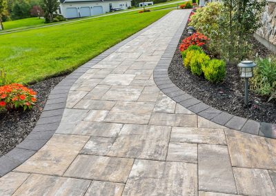 Wide stone walkway with dark edging, bordered by mulch, flowers, and green grass leading to a white house.