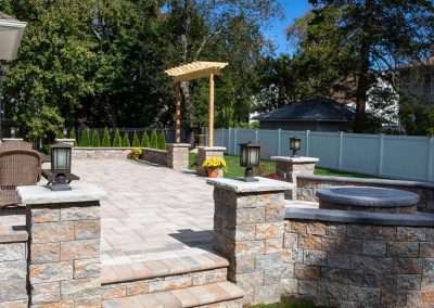Stone patio with steps, a fire pit, and pergola in a fenced backyard with outdoor seating and greenery.