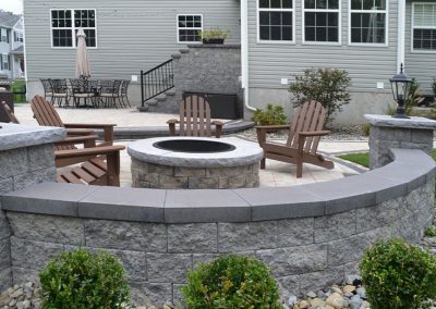 Stone fire pit with Adirondack chairs on a patio, surrounded by shrubs and a gray house in the background.