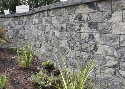 Gray stone retaining wall with a curved top, soil, and decorative plants in front, trees in the background.