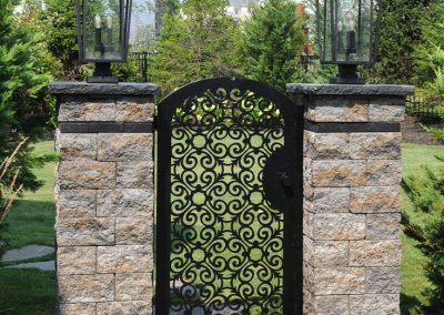 Ornate black metal gate between two stone pillars topped with lanterns, surrounded by greenery.
