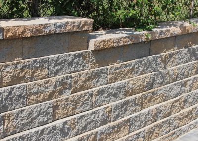 Stacked stone retaining wall with beige cap blocks, green vines, and a chain-link fence in the background.