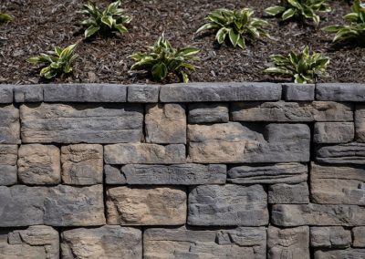 Gray stone retaining wall with mulch and small green plants growing on top.
