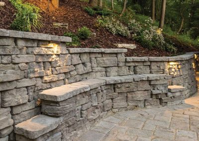 Stone retaining wall with built-in steps, illuminated by small lights, surrounded by trees and plants.