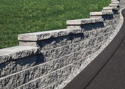 Stone retaining wall with tiered levels supports a grassy slope next to a paved driveway.