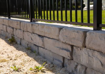 Stone retaining wall with a black metal fence on top, beside a grassy lawn and sandy soil with small weeds.