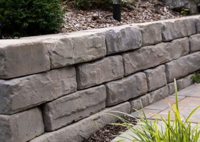 Short stone retaining wall with landscaping, plants, mulch, and a garden light along a pathway.