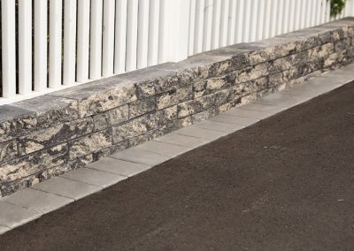 Stone retaining wall and gray pavers next to an asphalt road and a white vertical slat fence.
