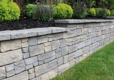 Gray stone retaining wall with landscaped plants and flowers on top, next to a green lawn.