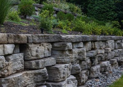 Stone retaining wall with garden beds and green plants above, bordered by decorative rocks at the base.