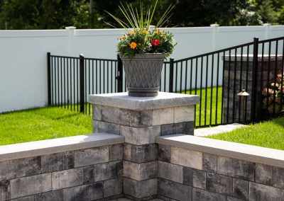 A gray planter with colorful flowers sits on a stone pillar in a paved backyard with a white fence.