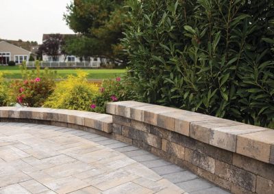 Curved stone patio wall with paving stones, lush green bushes, and grass in the background.